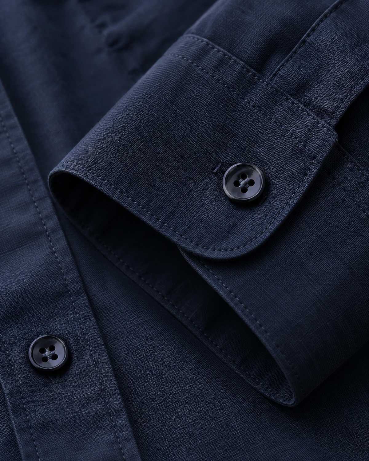 Primo piano del polsino di una camicia blu scuro, con bottone e cuciture visibili. / Close-up of a dark blue shirt cuff with a visible button and stitching.