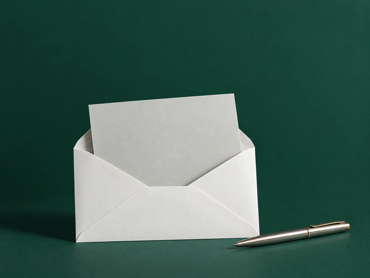 Busta bianca aperta con un foglio senza scritte e una penna su sfondo verde scuro. / An open white envelope with a blank sheet and a pen against a dark green background.
