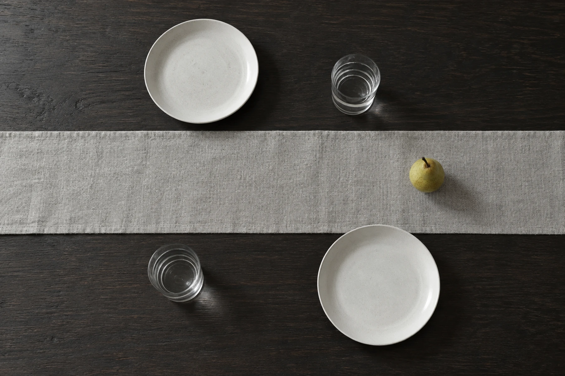Vista dall’alto di un tavolo scuro con due piatti bianchi, due bicchieri, un runner e una pera. / Overhead view of a dark table with two white plates, two glasses, a runner and a pear.
